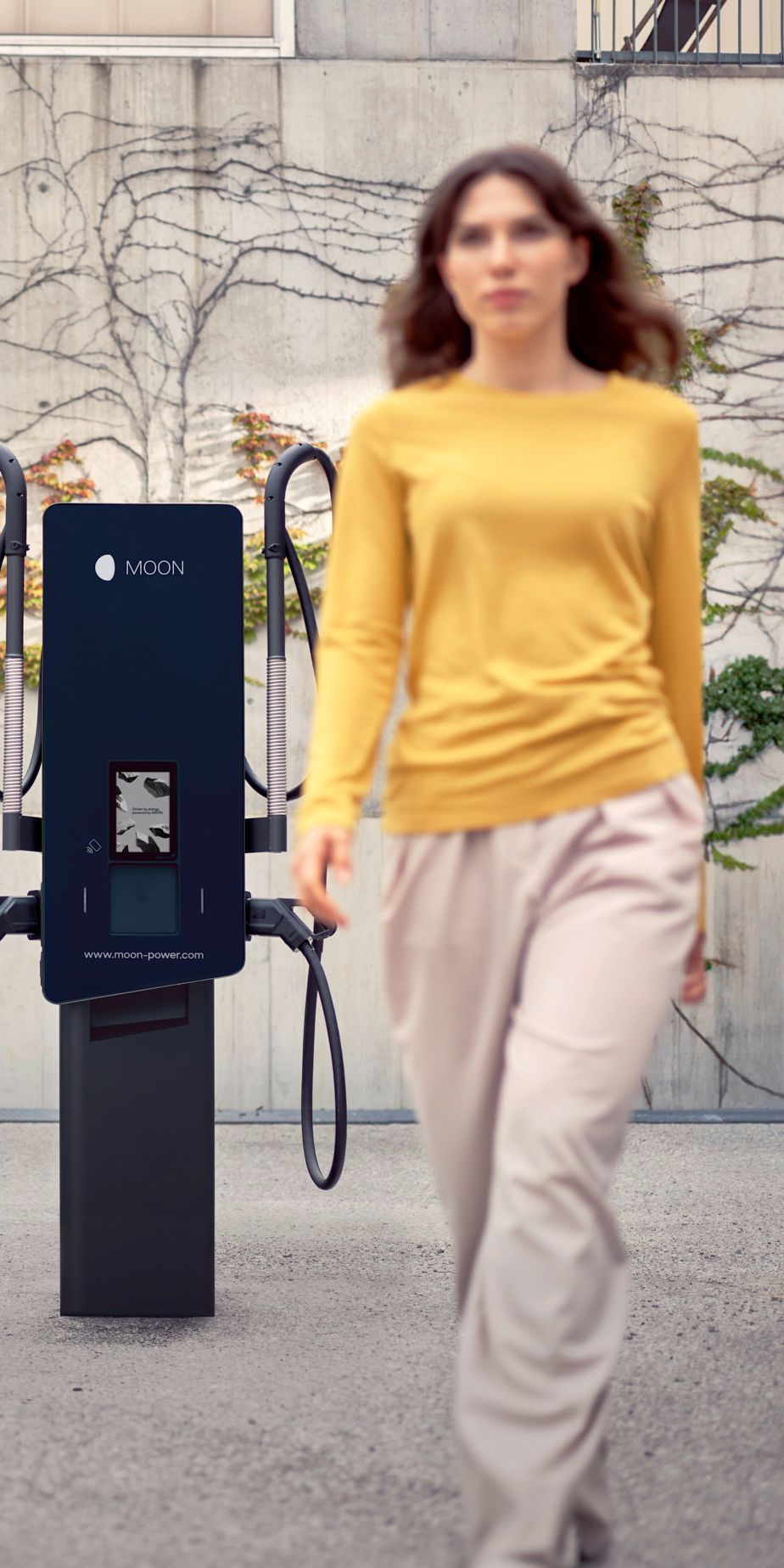 O femeie merge în fața unei stații de încărcare cu o mașină în fundal